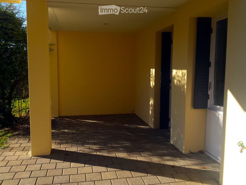 A covered outdoor area with yellow walls, brick flooring, and a view of greenery outside.