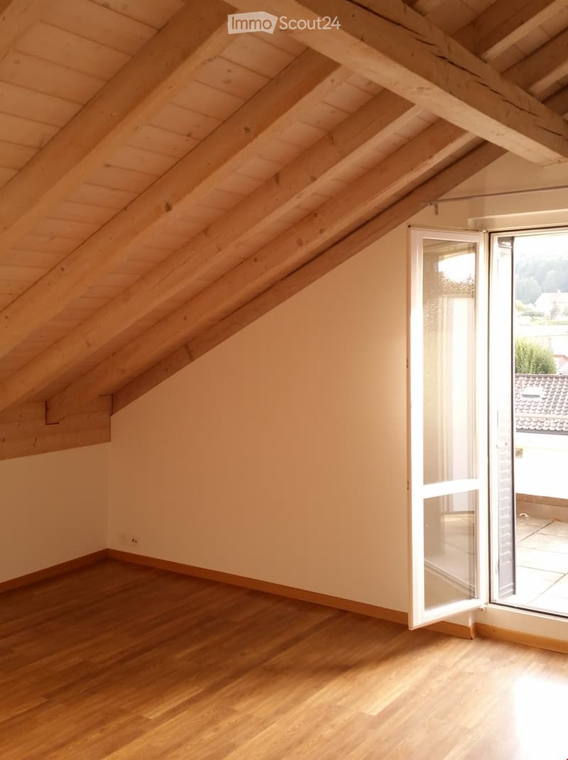 An empty attic room with wooden floors and exposed wooden beams on the ceiling. A large window with white frames is open, allowing natural light to enter.