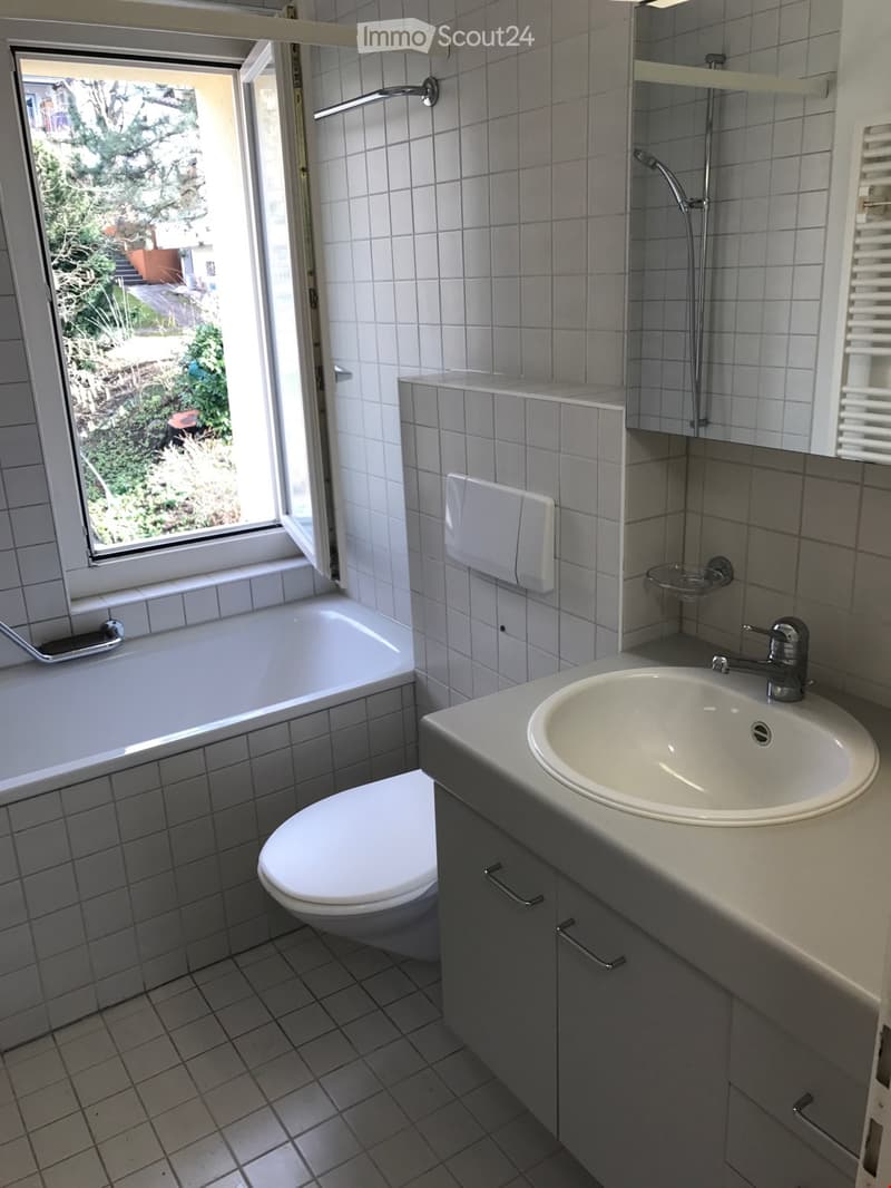 Modern bathroom with white tiles, bathtub, toilet, sink, and showerhead. Features a large window with a view of greenery outside.