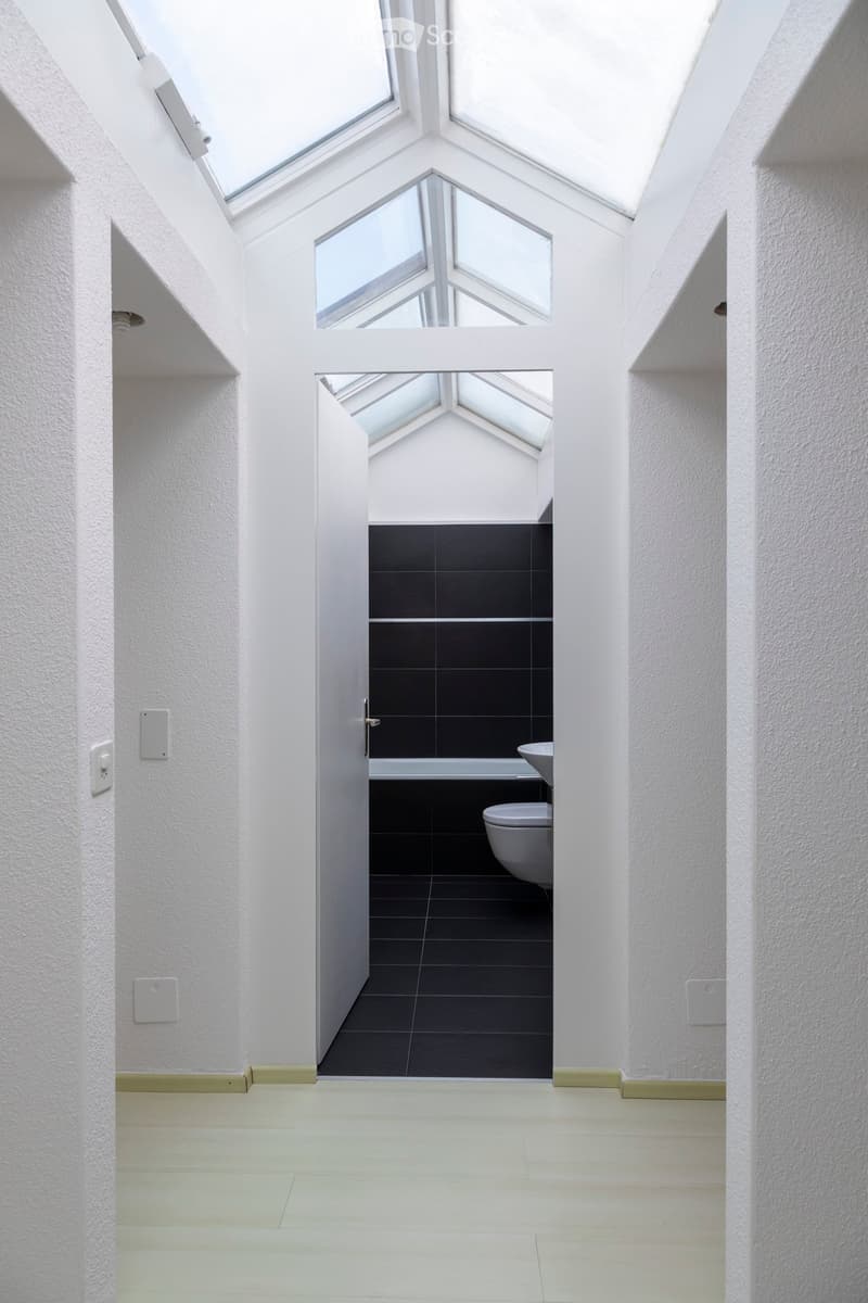 Bright, minimalist hallway with white walls and light-colored flooring, leading to an open bathroom with dark tiles and modern fixtures.