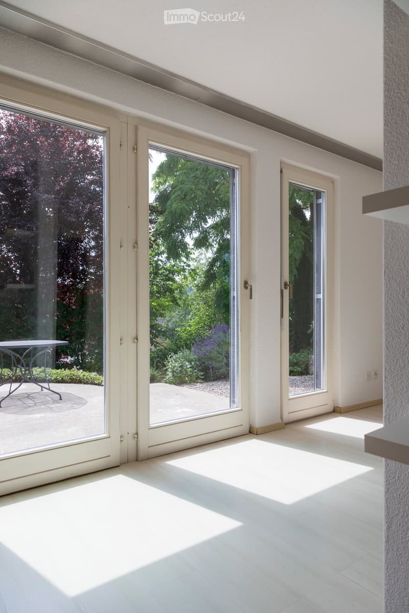 Bright, empty room with large glass doors leading to a garden, white tiled floor, and modern white walls.