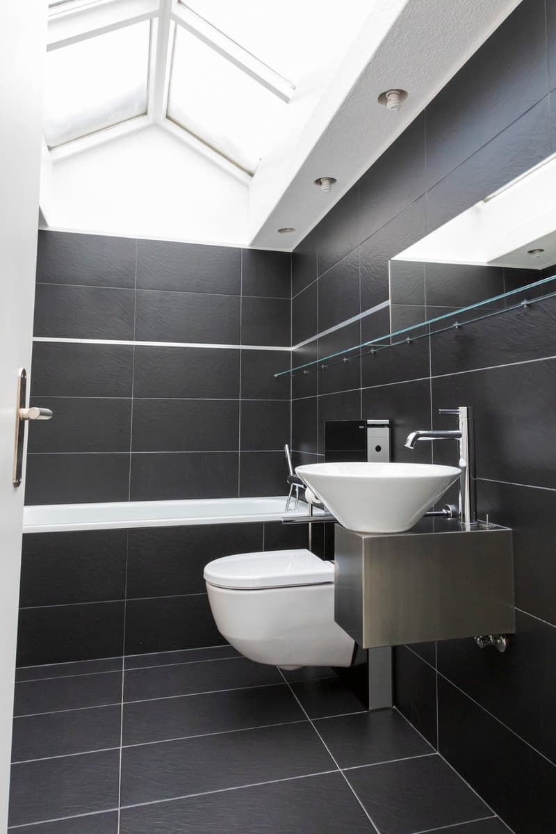 Modern bathroom with black tiles, white fixtures, and a skylight window above the bathtub.