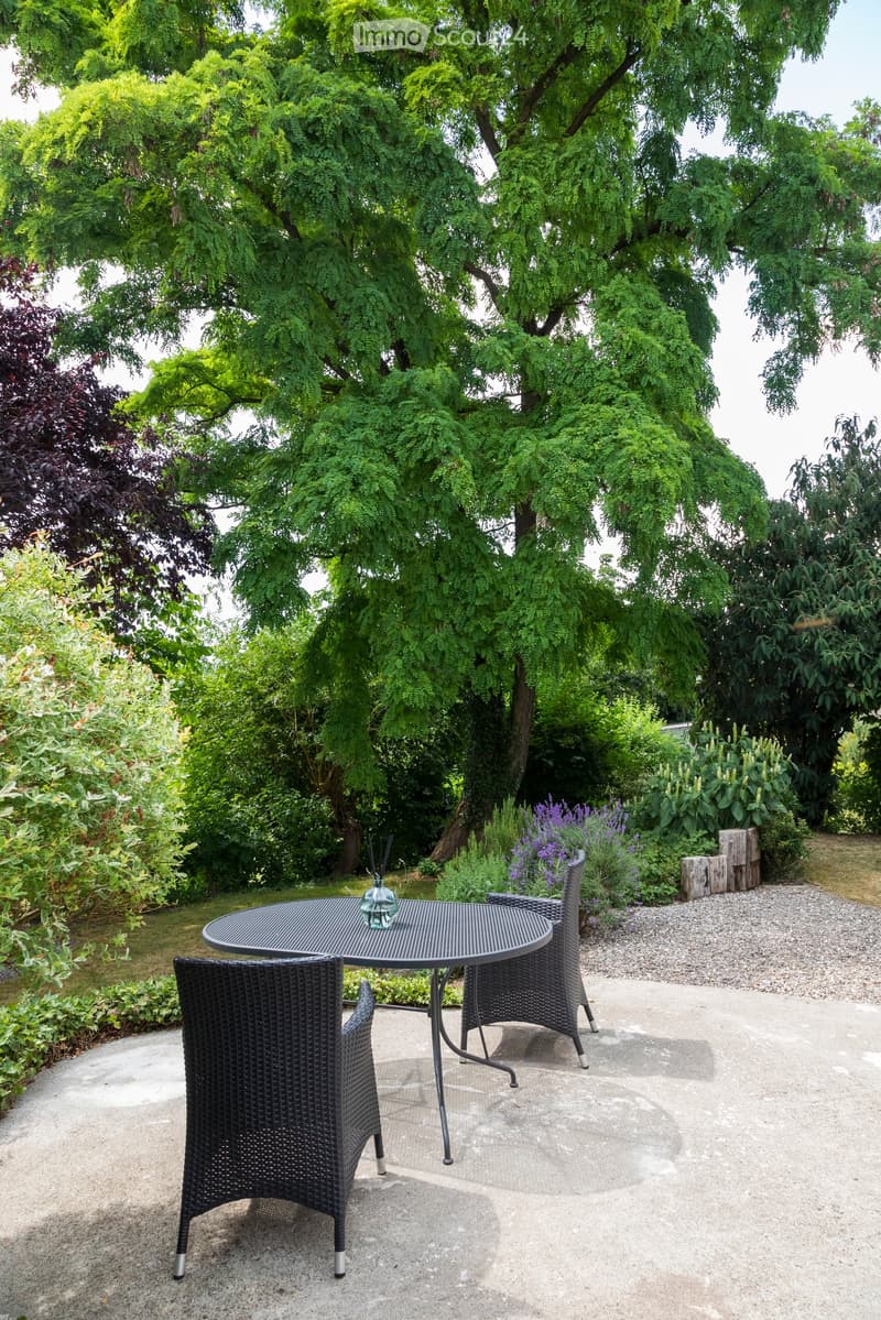 Garden area with a round metal table and two wicker chairs, surrounded by lush greenery and flowers.