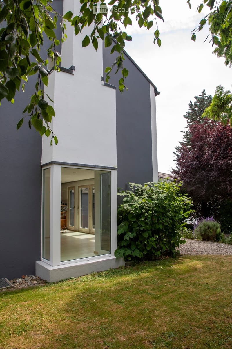 A modern house with a raised ground floor, featuring a large glass extension leading to a well-maintained garden with green grass, bushes, and trees.