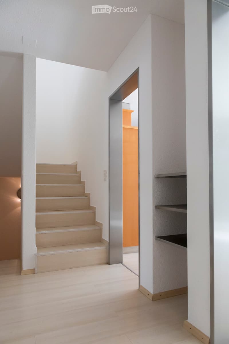 A modern, minimalist hallway with light wooden flooring, white walls, and a staircase leading upwards. A small built-in shelf is visible on the right side, and an open doorway leads to another room with orange walls.