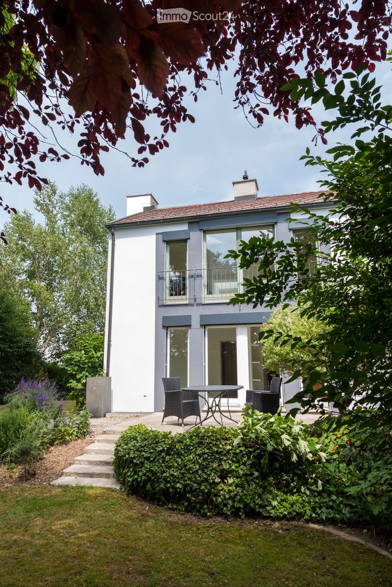 A modern two-story house with a balcony and large windows, surrounded by a well-maintained garden with a patio area featuring a table and chairs.