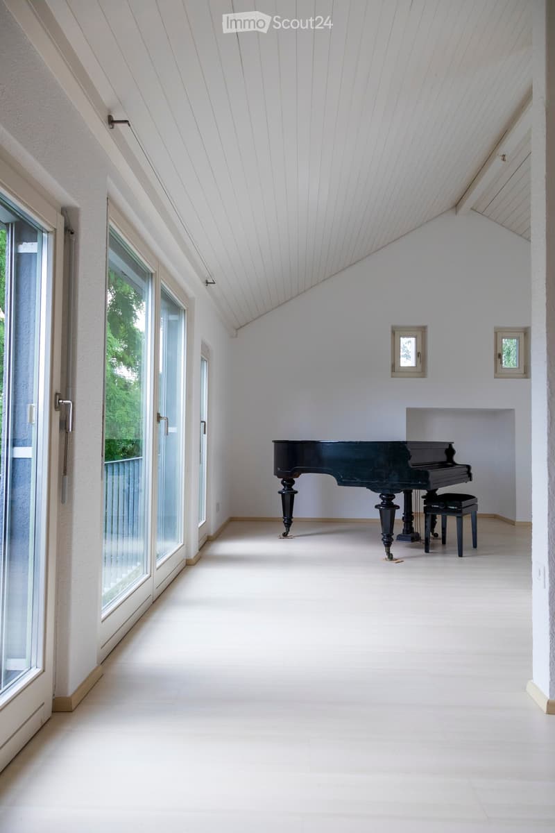Bright, spacious room with white walls and a high, slanted ceiling. Features a grand piano and a matching bench, large windows with views of greenery, and a clean, minimalist design.