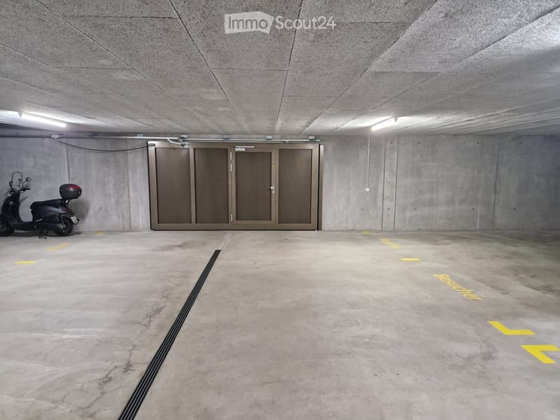 Underground parking area with concrete walls and floor, a single scooter parked on the left, and a large garage door in the center.