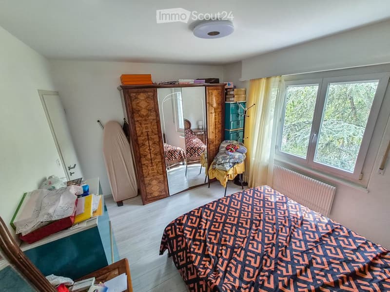 Bedroom with a bed, wardrobe, mirror, chair, table, and window with curtains. The room has wooden flooring and a radiator under the window.