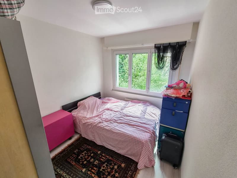 Cozy bedroom with a bed covered in a pink and white patterned duvet, a nightstand, a suitcase, and a colorful rug on the floor.