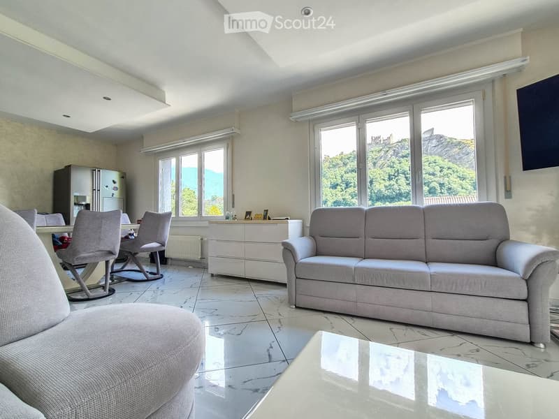 Modern living room with large windows offering a scenic view, featuring a grey sofa, ottoman, coffee table, dining area with chairs, and a refrigerator in the background.
