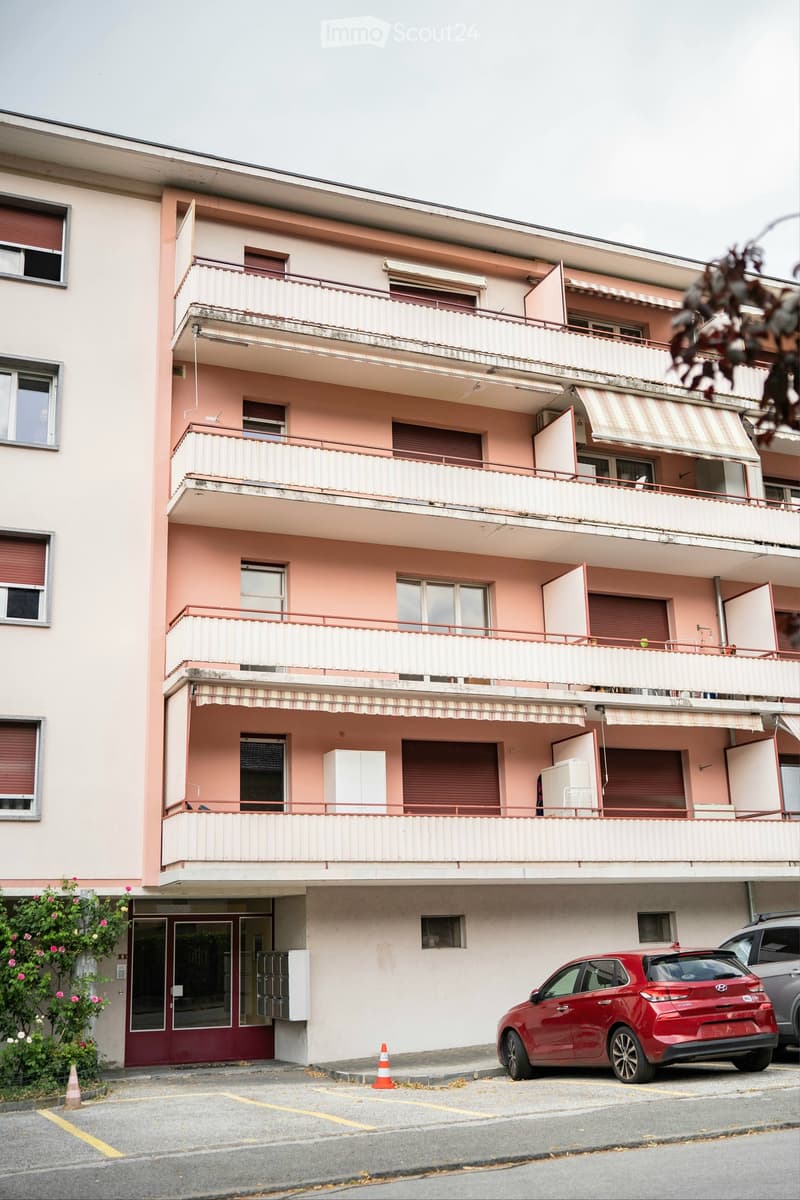 Four-story apartment building with pink facade, multiple balconies, and striped awnings. A red car is parked in the driveway.