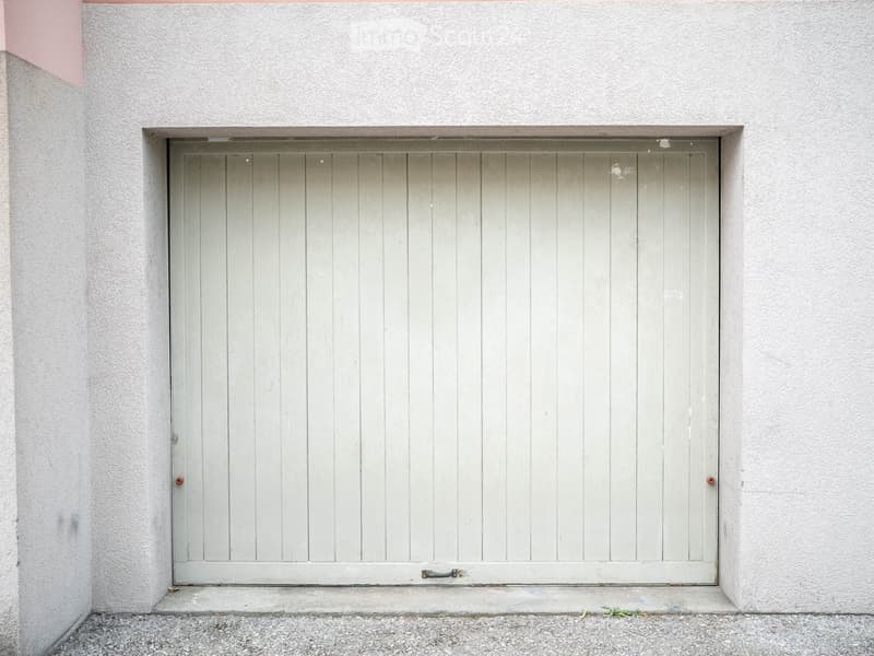 A closed garage door with a light grey finish, set within a white textured wall. The door features vertical slats and a simple handle.