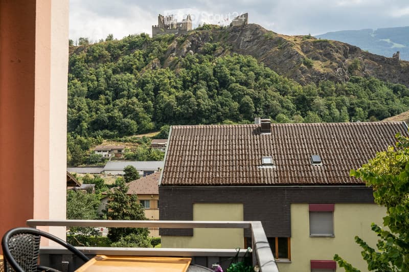 Balcony overlooking a town with a castle on a hill in the background, featuring a wooden table and a wicker chair.