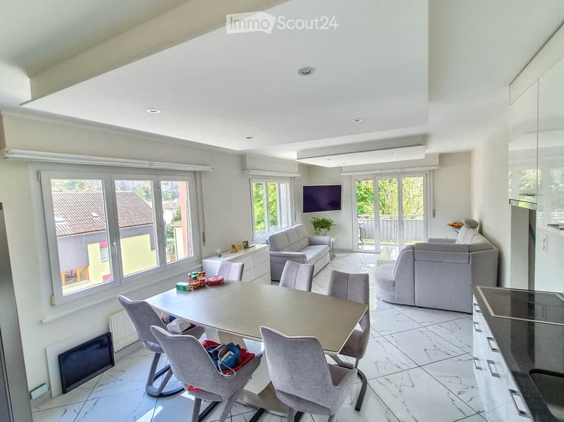 Bright living room with large windows, white walls, and a high ceiling. Features a sofa, chairs, a dining table, and a television. The room has a modern design with marble flooring and ample natural light.
