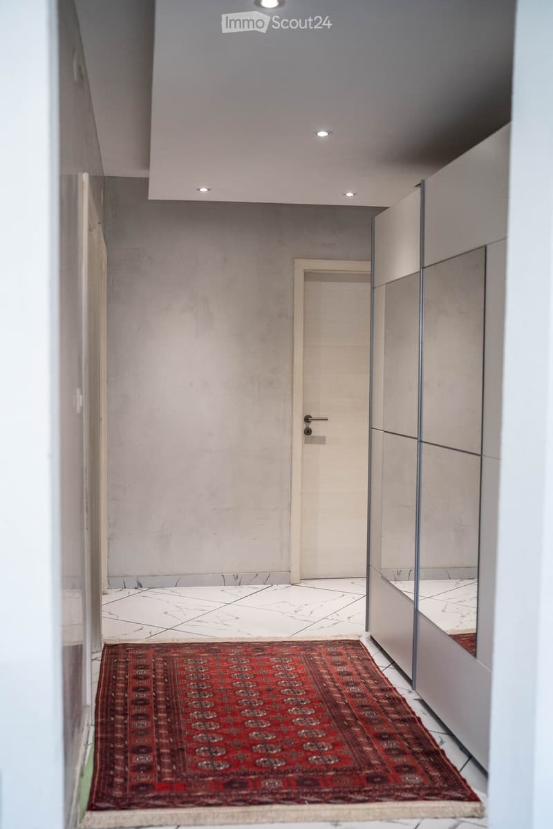 Modern hallway with white tiled floor, grey walls, recessed ceiling lights, and a red patterned rug placed on the floor.