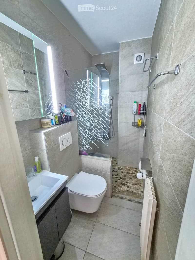 Modern bathroom with a white sink, toilet, glass-enclosed shower, and a radiator. The walls and floor are tiled in a light, neutral color, and the shower features a decorative glass panel.