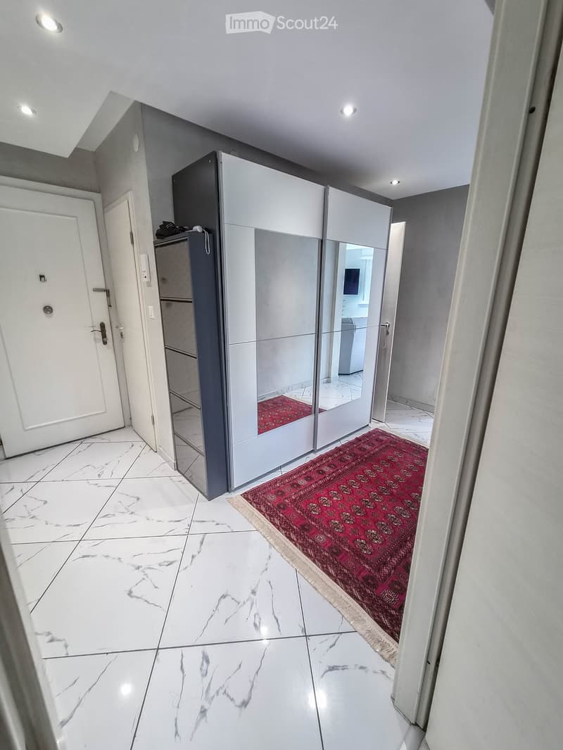 Modern hallway with white marble flooring, white walls, recessed lighting, and a red patterned rug leading to a mirrored closet with glass doors.