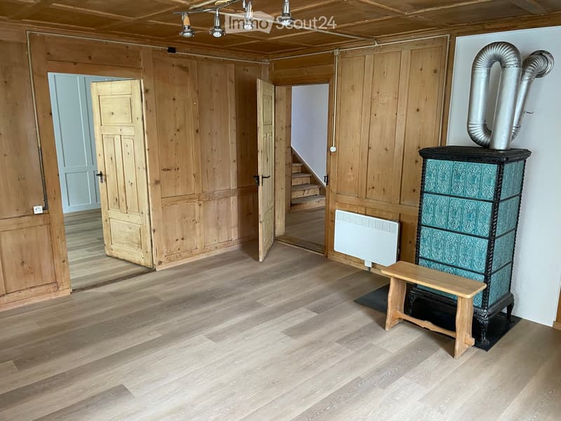 Empty room with wooden walls, wooden floor, a staircase leading upwards, a radiator, and a decorative stove with a bench in front of it.