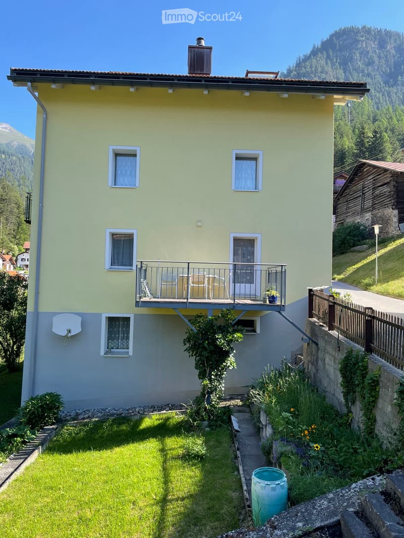 Two-story house with yellow facade, white trim, and balcony on the upper level. The house features multiple windows, a small garden, and is situated in a scenic mountainous area.