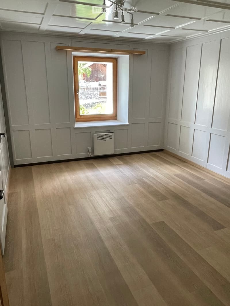 Empty room with light wooden flooring, white walls, and a large window with wooden frame. A small white radiator is visible below the window.