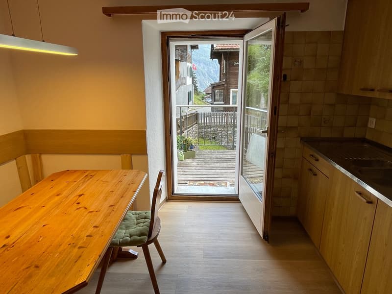 Bright room with wooden floor, large open door leading to a balcony with a scenic mountain view, a wooden dining table with a single chair, and modern kitchen cabinets.
