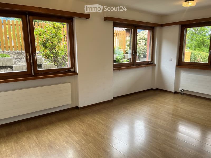 Empty room with wooden flooring, white walls, and large windows with wooden frames, offering a view of greenery outside. Radiators are installed below the windows.
