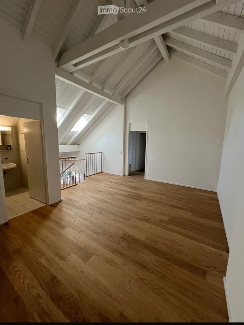 Empty attic space with wooden flooring, white walls, exposed wooden beams, and skylights providing natural light. Features a staircase leading to another area.