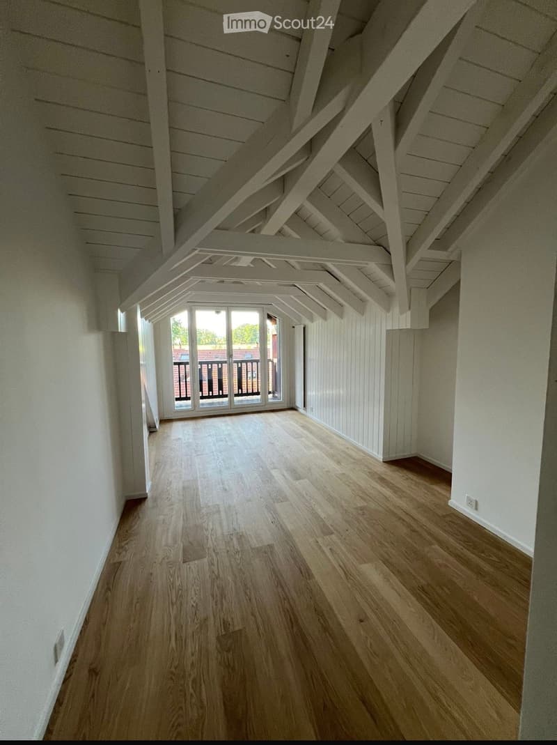 Empty attic space with wooden flooring, white walls, and exposed wooden beams. Features a large window with a view of a balcony and outdoor area.