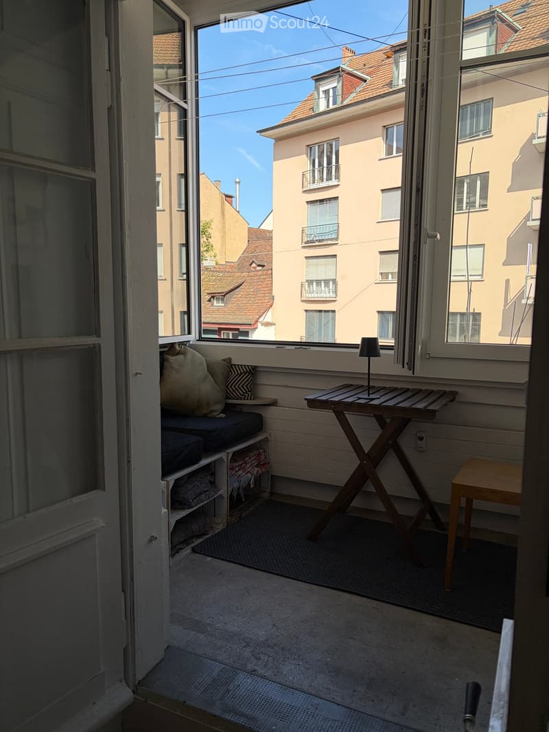 Balcony with wooden table, stool, bench, and lamp, overlooking a cityscape with buildings and clear sky.