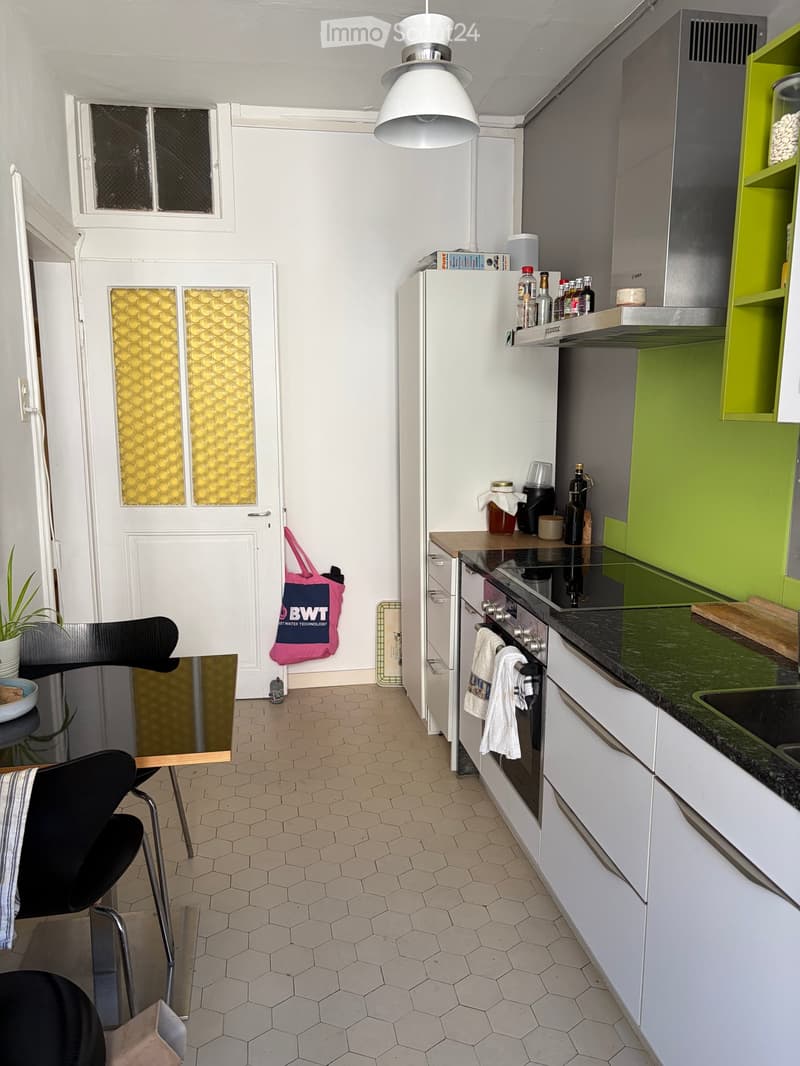 Modern kitchen with white cabinets, green accents, and stainless steel appliances. Features a hexagonal tiled floor, a black chair, a small table, and a handbag hanging on the door.