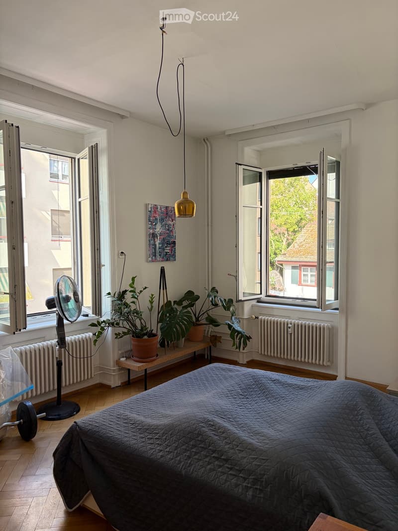 Bright bedroom with wooden flooring, large windows, a bed with a dark quilt, a fan, potted plants on a small table, and a painting on the wall.
