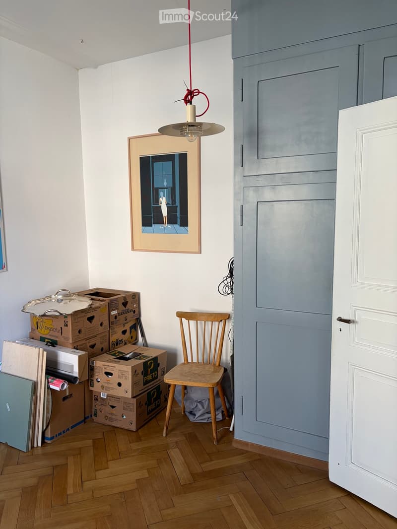 A room with wooden flooring, a wooden chair, a picture frame on the wall, a pendant light, and several cardboard boxes stacked near the wall.