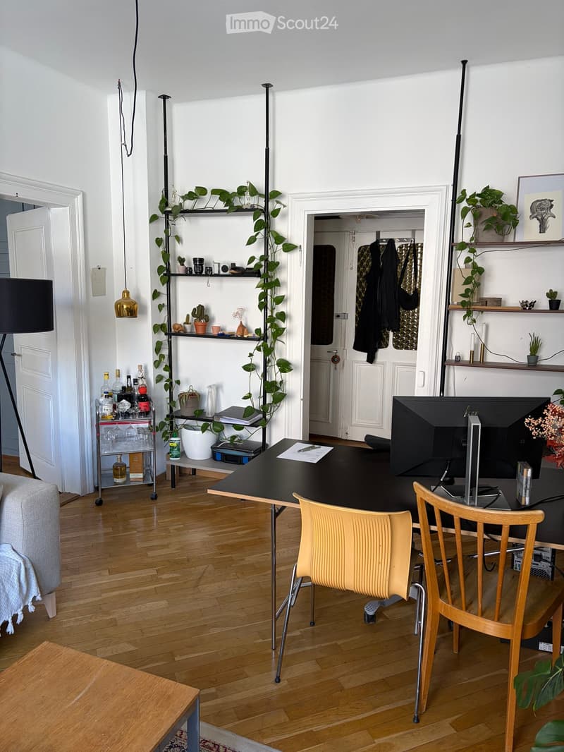 A modern, well-lit room with wooden flooring, a large desk with a screen, two chairs, a sofa, a shelf with plants and decorative items, and a small bar cart with bottles and glasses.
