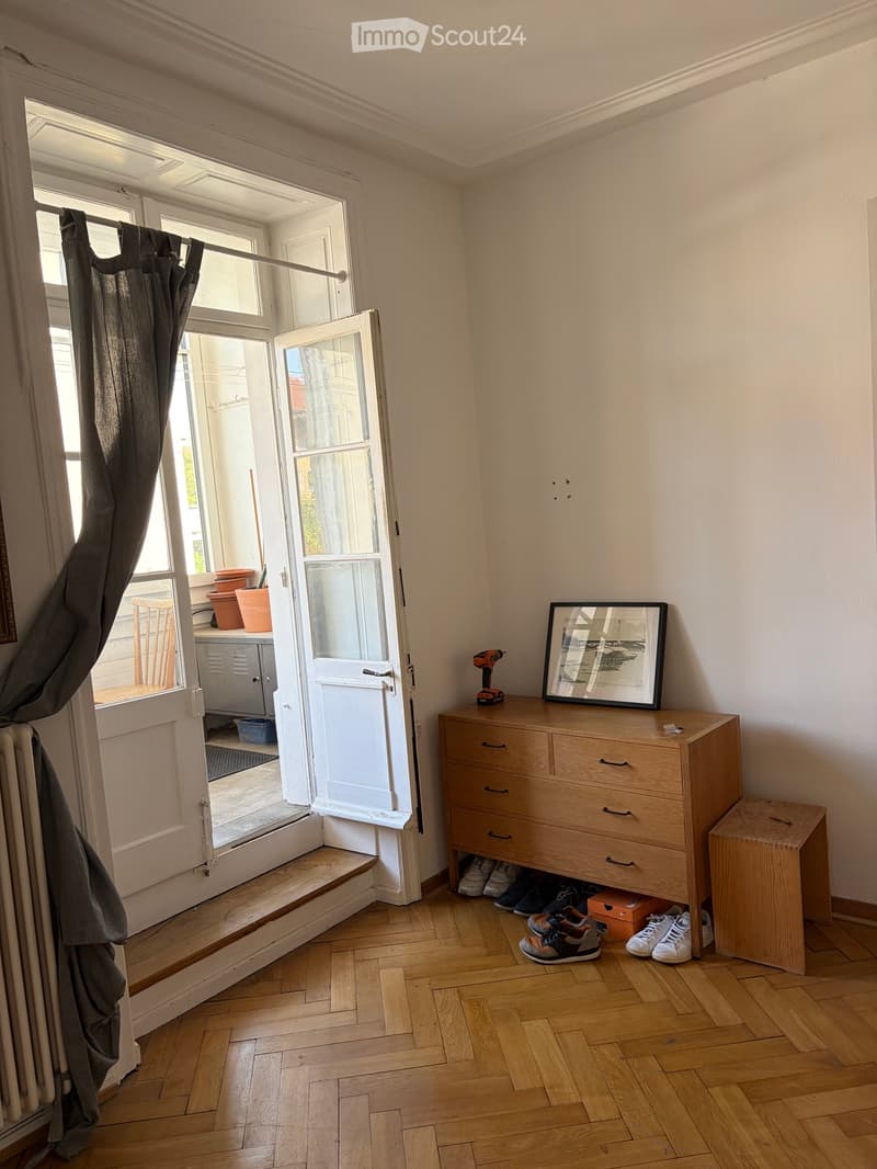 A small, well-lit room with wooden flooring and white walls. Features include a chest of drawers with a picture frame and drill on top, a curtained window leading to a small outdoor area with potted plants, and several pairs of shoes placed near the chest