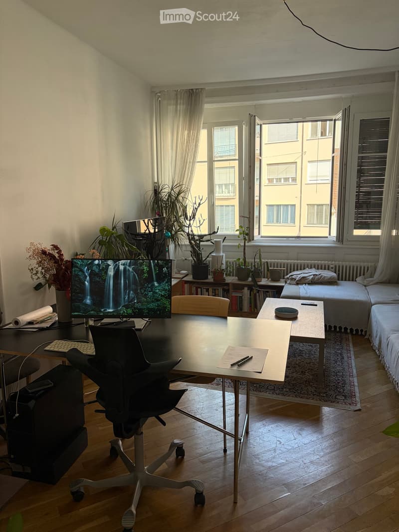 A well-lit room with a desk, ergonomic chair, laptop, and a TV screen displaying a waterfall. There is a small sofa and a coffee table with a remote control and a plate. The room has wooden flooring and large windows with curtains, allowing natural light