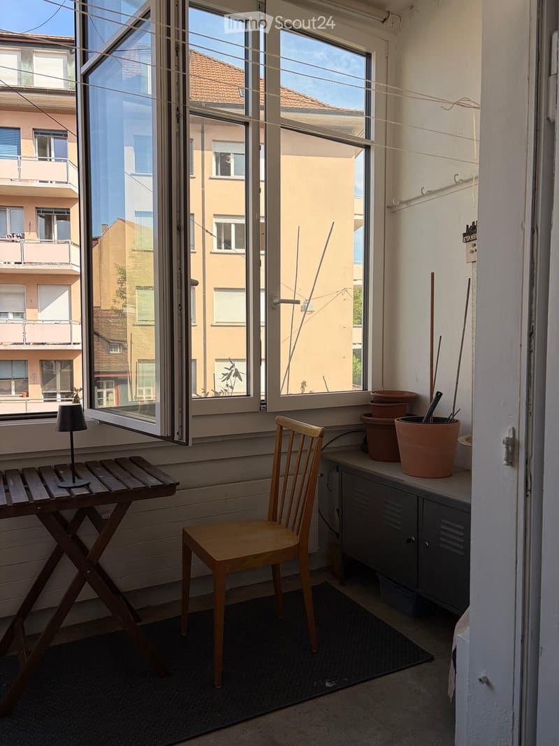 Balcony with wooden chair, small table, lamp, and several pots on the windowsill. Large windows open to a view of neighboring buildings.