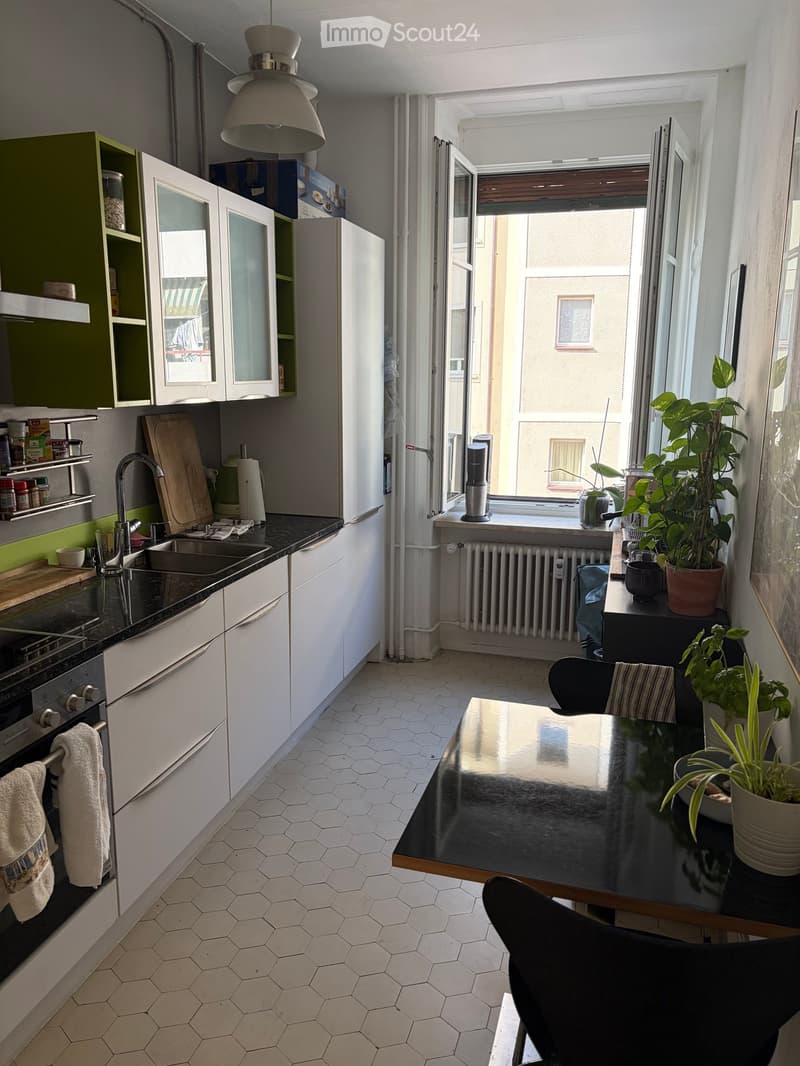 A modern kitchen with white cabinets, a black countertop, and a dining area with a black table and chairs. The kitchen features a sink, oven, and various utensils. A potted plant is placed on the windowsill, and the room has a view of a building outside.