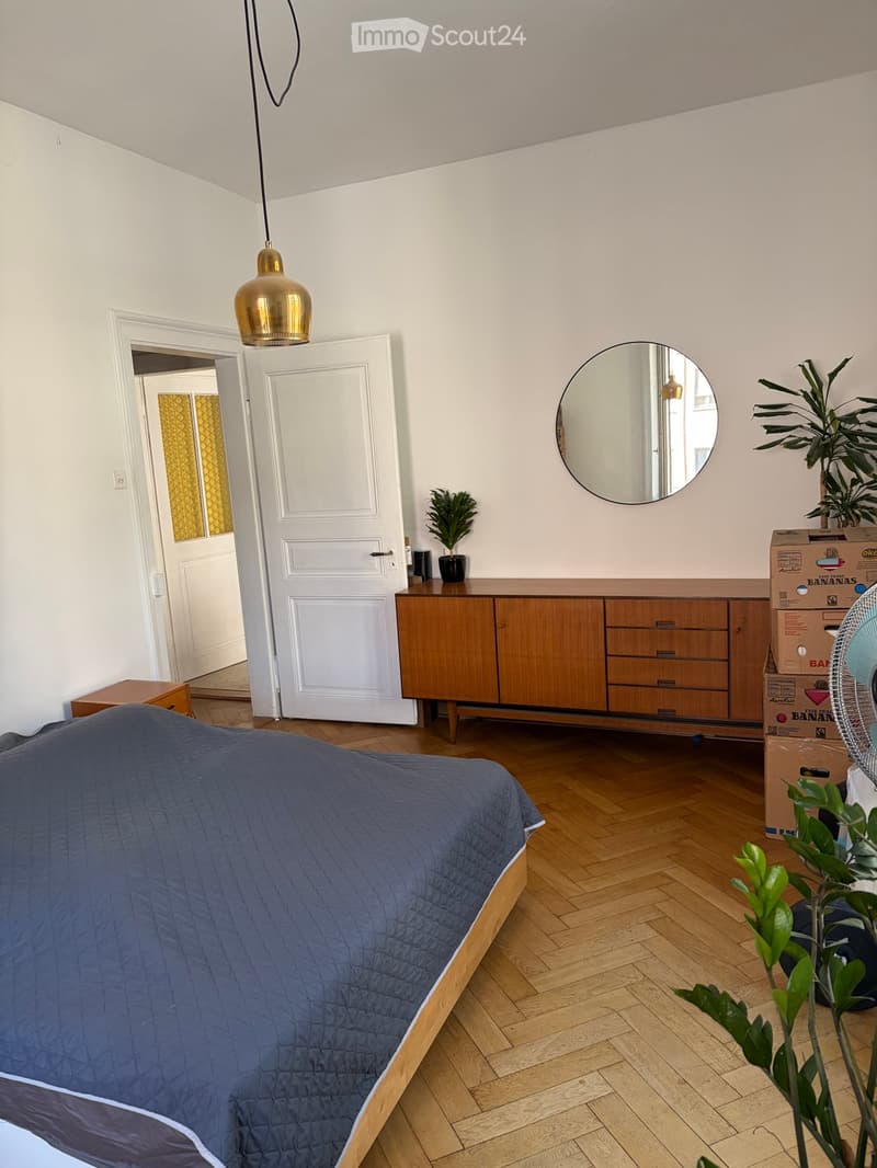 A modern bedroom with a wooden bed, a blue quilt, a wooden cabinet, a round mirror, a fan, and potted plants.