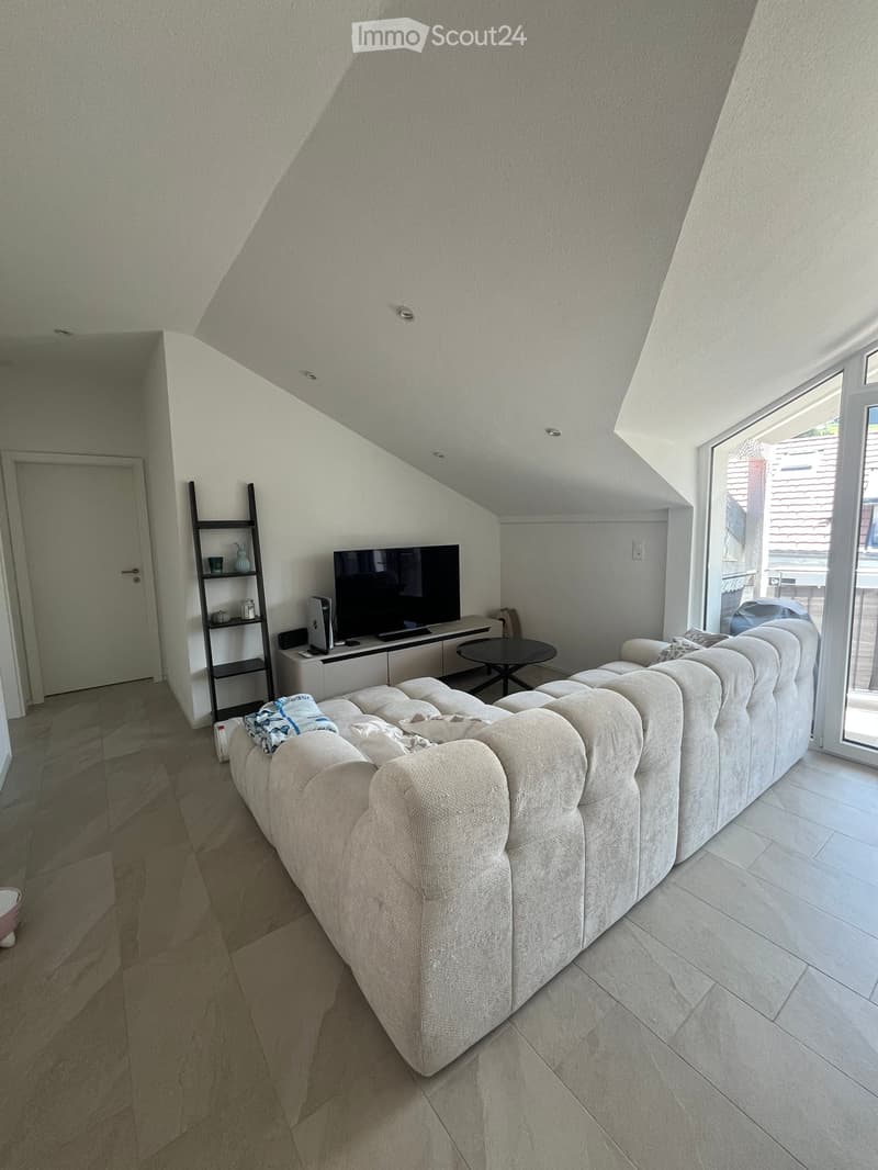 Modern living room with a large sectional sofa, a small round table, a TV stand with a television, a shelf with decorative items, and a laptop on the TV stand. The room has a slanted ceiling and large windows letting in natural light.
