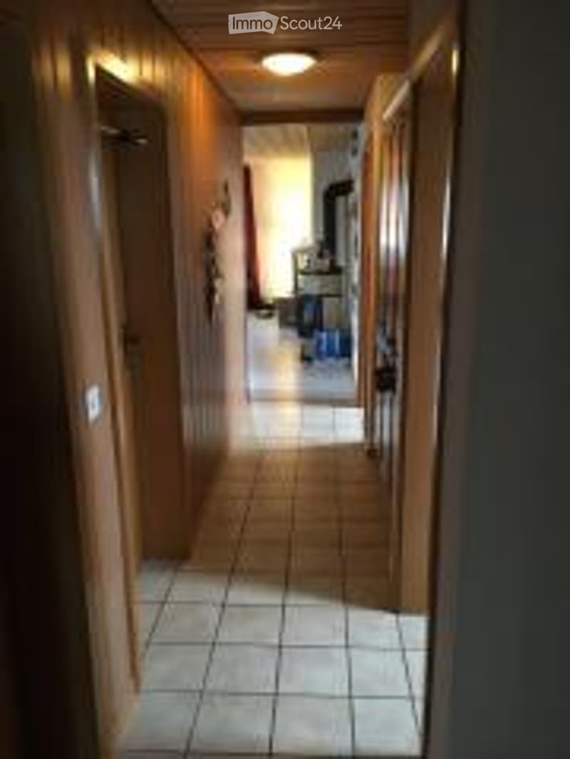 A narrow hallway with white tiled flooring, wooden walls, and a recessed ceiling light. The hallway leads to an open area with visible furniture.
