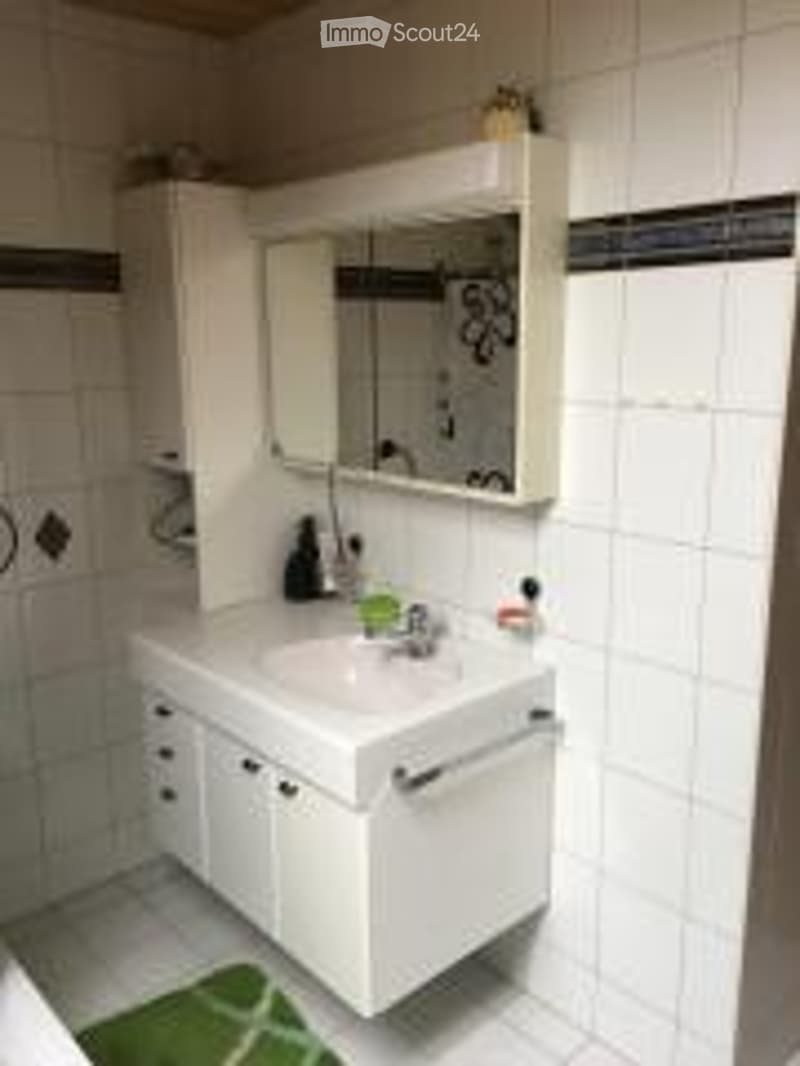 White bathroom with a sink, mirror, and cabinet. The walls are tiled, and a small green rug is placed on the floor.
