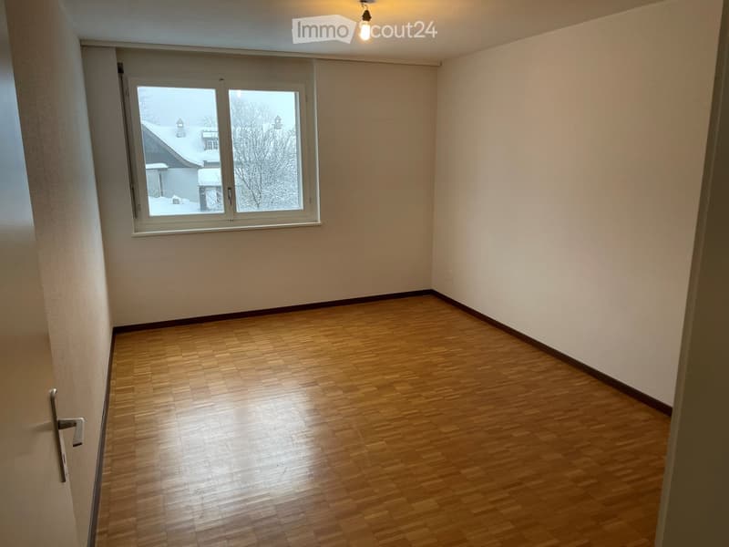Empty room with wooden flooring, white walls, and a large window showing a snowy outdoor scene.