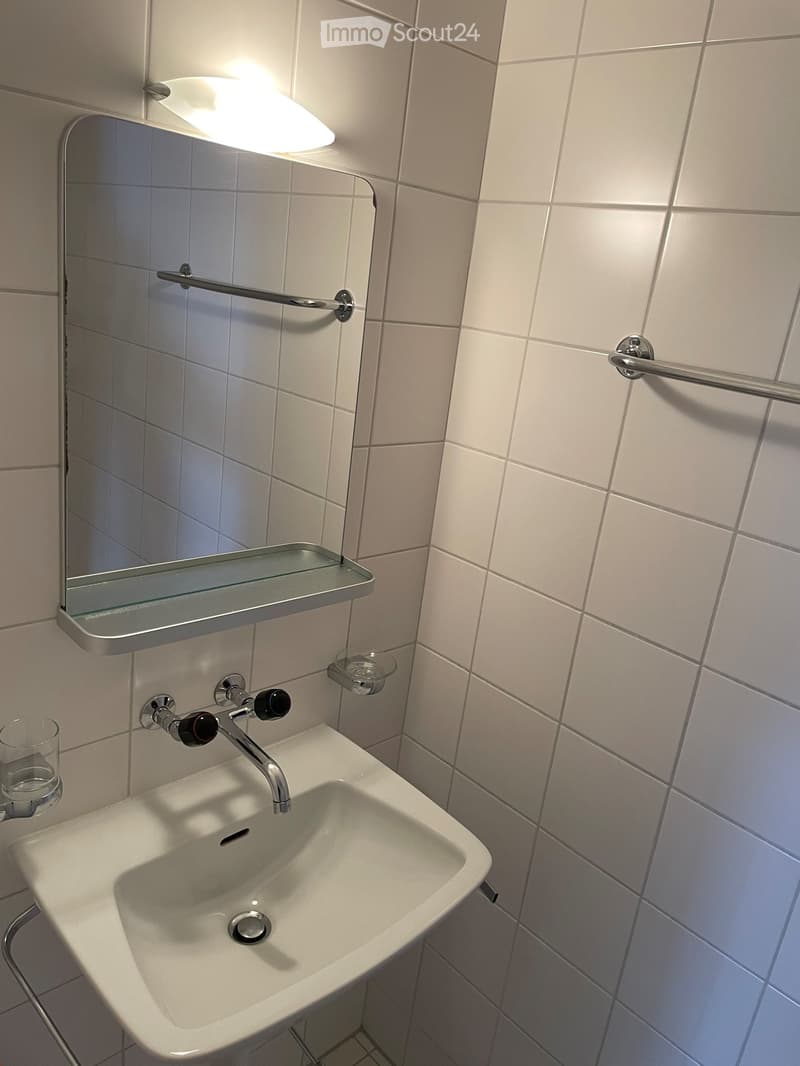 Modern bathroom sink with a chrome faucet, a built-in mirror with a glass shelf above it, and a small glass cup on the left side.