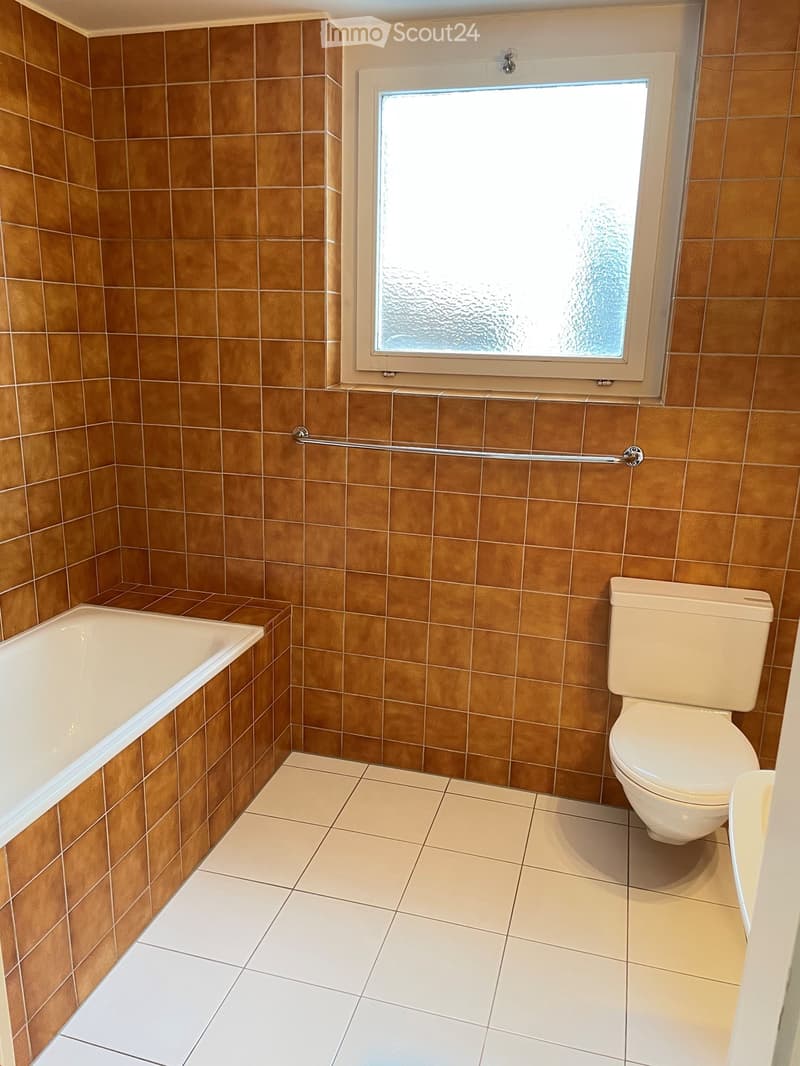 Bathroom with brown tiled walls, white bathtub, toilet, and a handrail near the window.