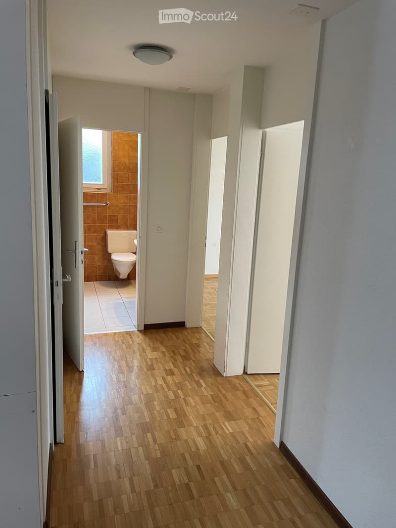 A clean, well-lit hallway with wooden flooring and white walls, leading to a bathroom with a toilet and tiled walls.