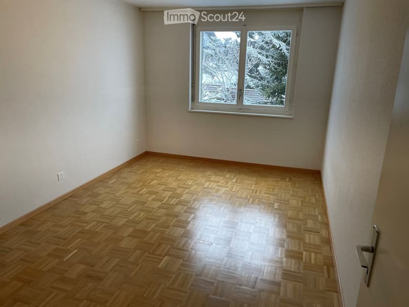 Empty room with wooden parquet flooring, white walls, and a large window showing a snowy outdoor scene.