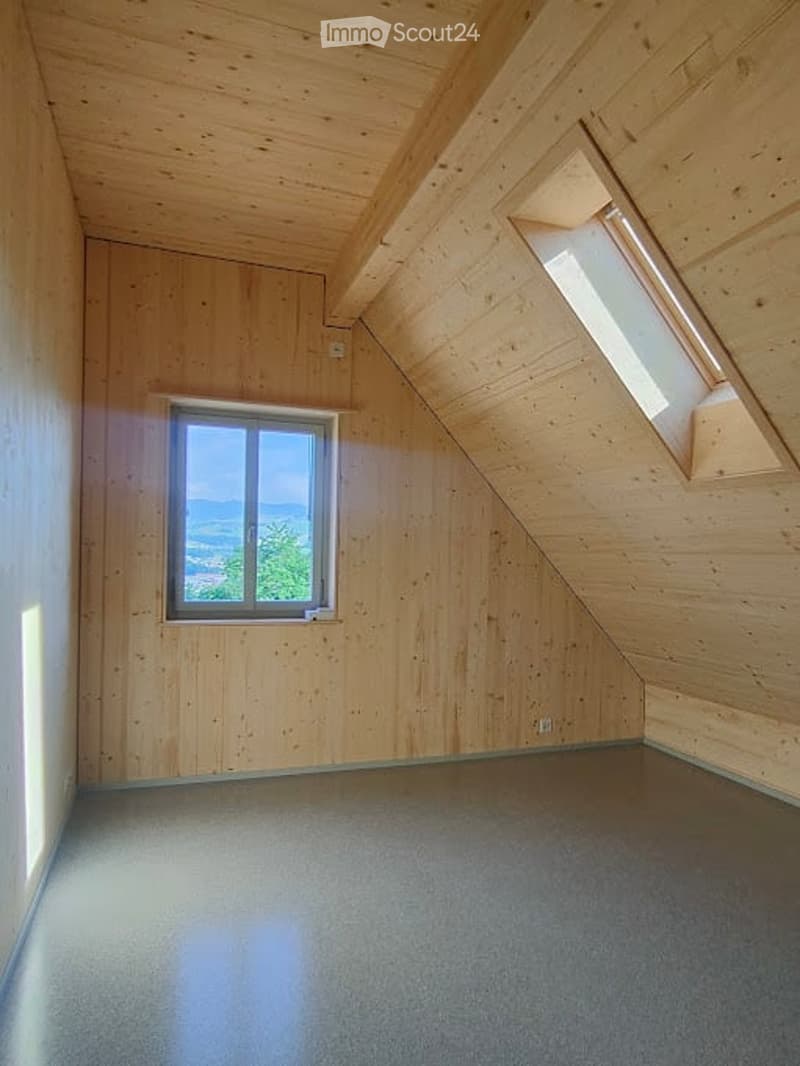 Empty attic room with wooden walls and ceiling, two windows, and a sloped roof.