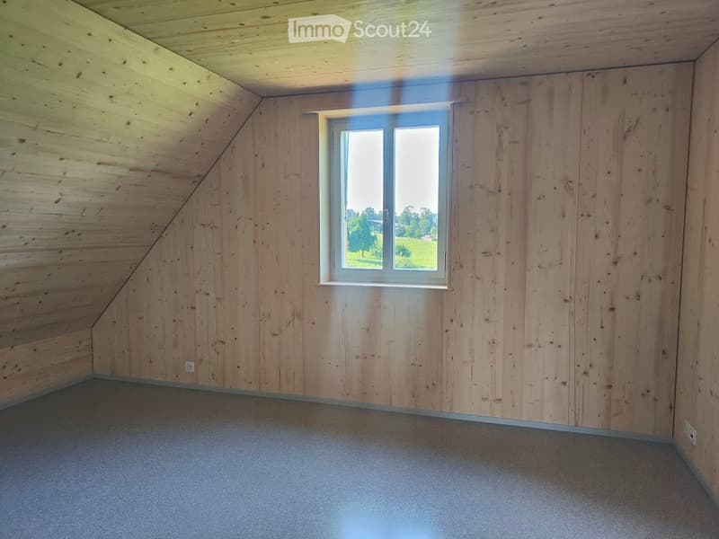 Empty attic room with wooden walls and ceiling, carpeted floor, and a single window offering a view of the outdoors.