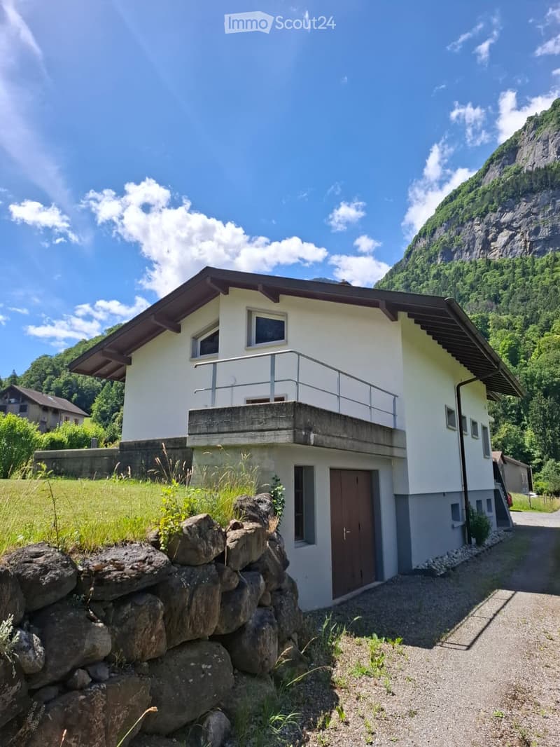Two-story house with white walls, dark roof, and balcony overlooking a mountainous landscape.